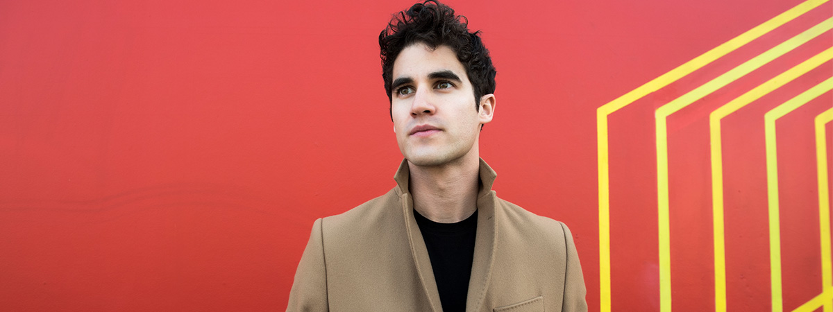 Darren Criss standing in front of a red and yellow geometric background
