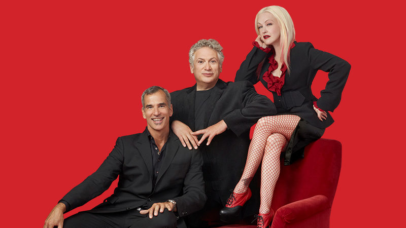 Jerry Mitchell, Harvey Fierstein, and Cyndi Lauper sitting on a chair in front of a bright red background.