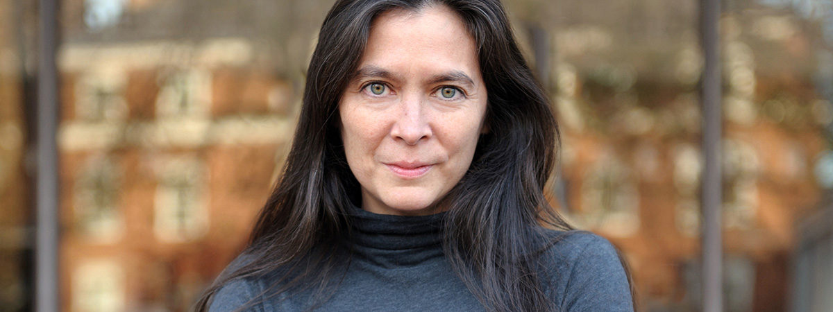 Diane Paulus wearing a gray turtleneck in front of a reflective surface in a metropolitan area.