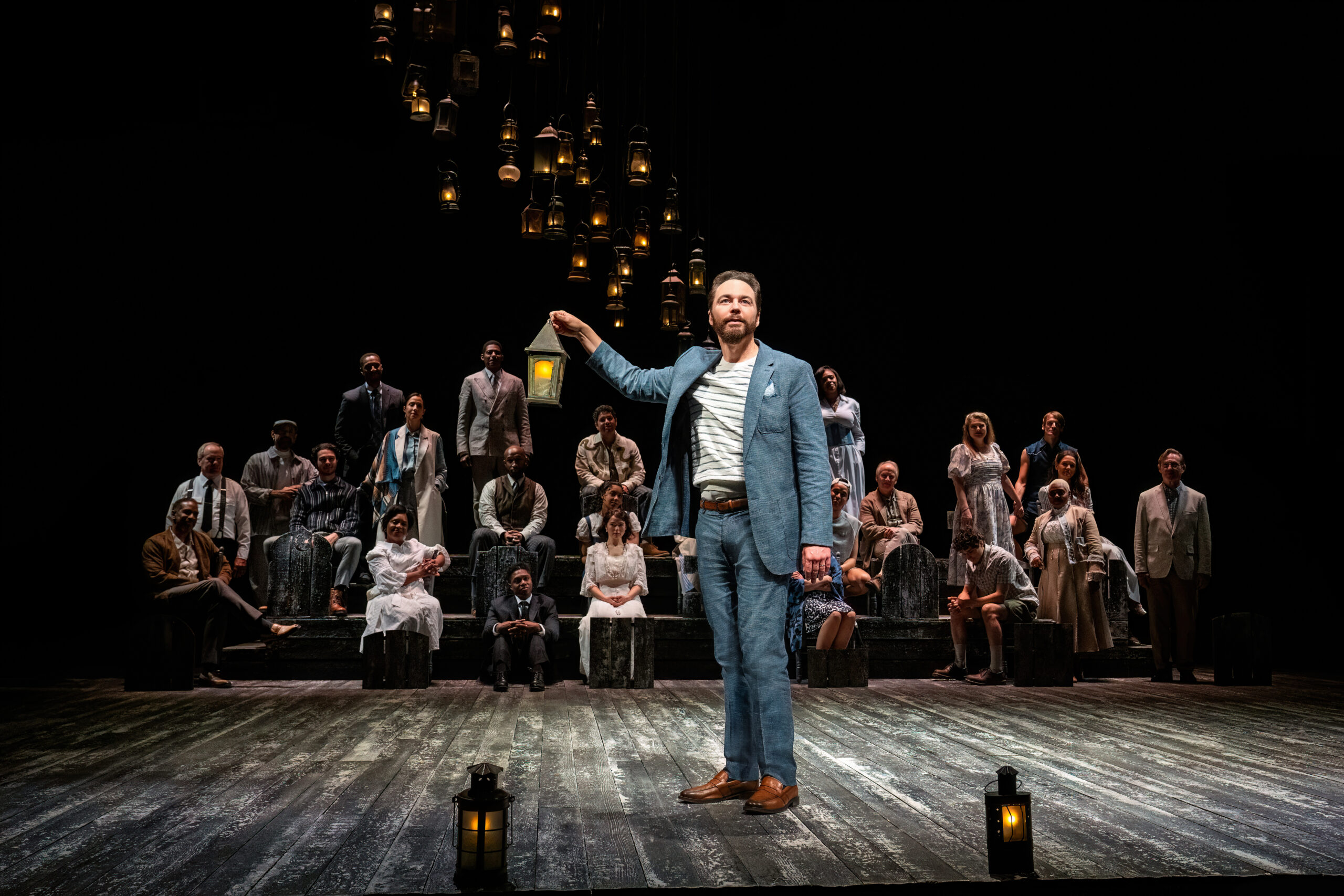 A character holding a lamp downstage with the cast behind sitting upstage on risers