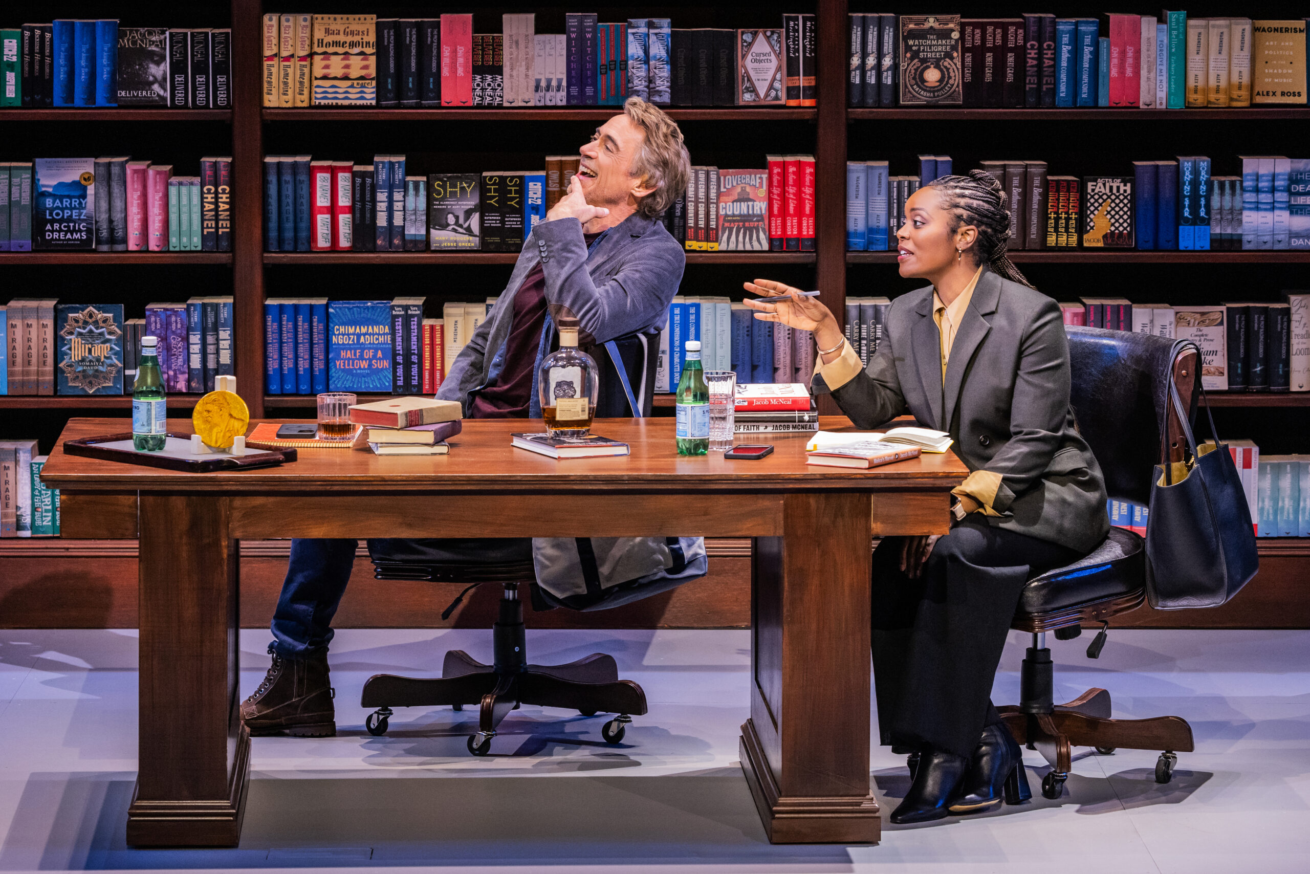 Two characters sitting at a desk looking to the left with a bookshelf behind them