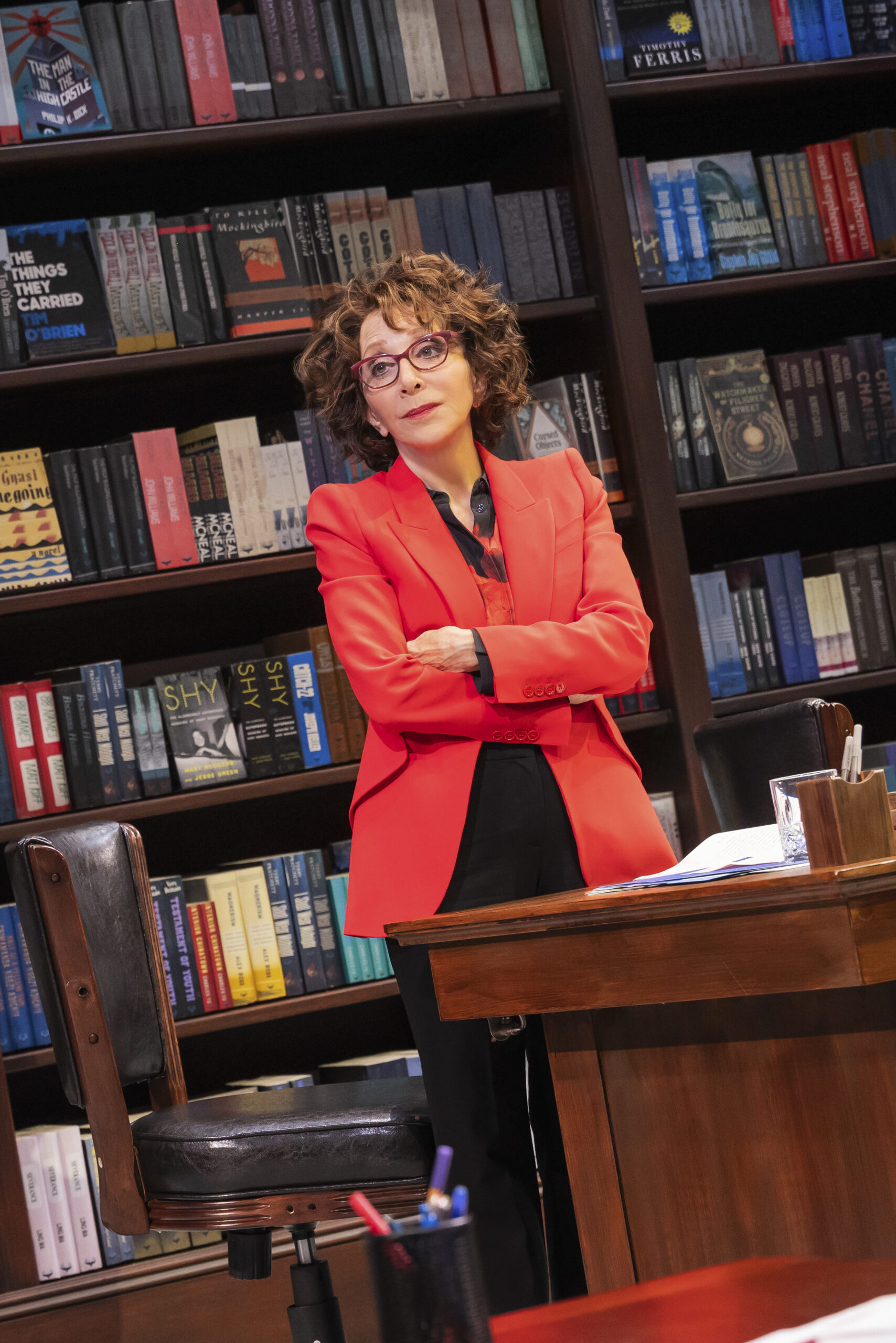 Character on stage in red blazer with bookshelf behind them.
