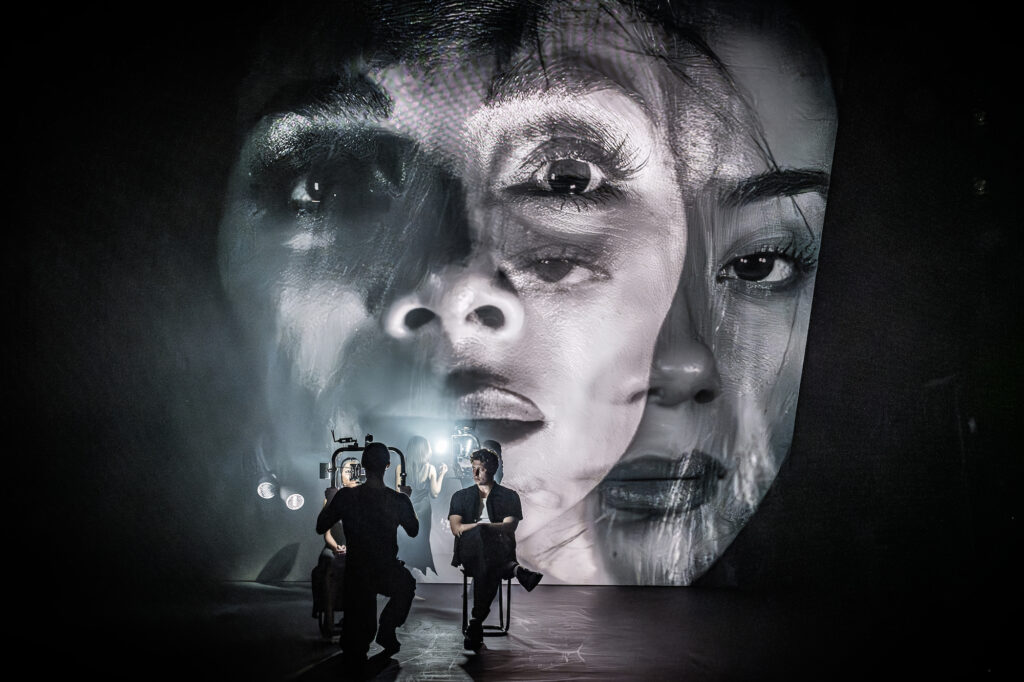 Nicole Scherzinger and Hannah Yun Chamberlain on screen with Tom Francis seated in Sunset Blvd. Photo by Marc Brenner.